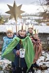 Vier Kinder mit goldenen Kronen und bunten Umhängen stehen im Schnee, eines hält einen großen goldenen Stern auf einer Stange und eine Spendendose